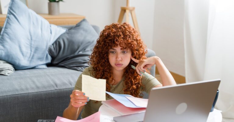 Young woman frowning while managing her finances and bills, surrounded by paperwork and using a laptop