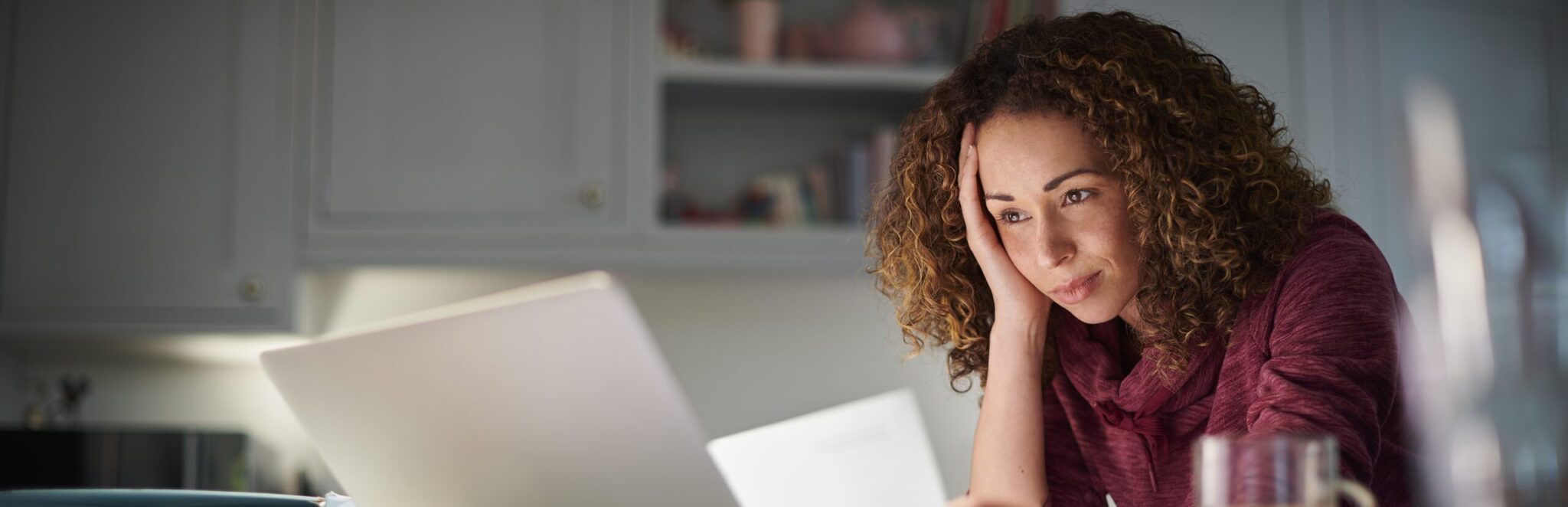 Woman in her kitchen in the evening with a cup of coffee looking at her laptop and the document she has in front of her