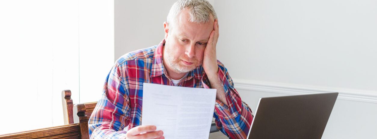 Worried mature man in front of a laptop looking at a bill in his hand while sitting at a table