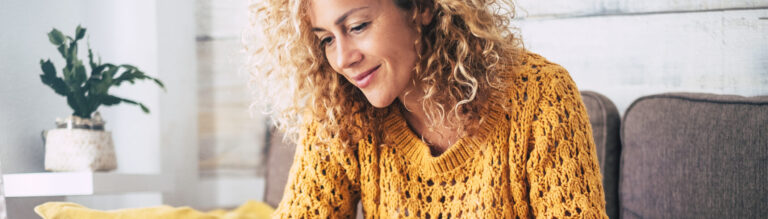 Middle aged lady with blonde curly hair and an burnt yellow sweater works at the her computer on the sofa at home