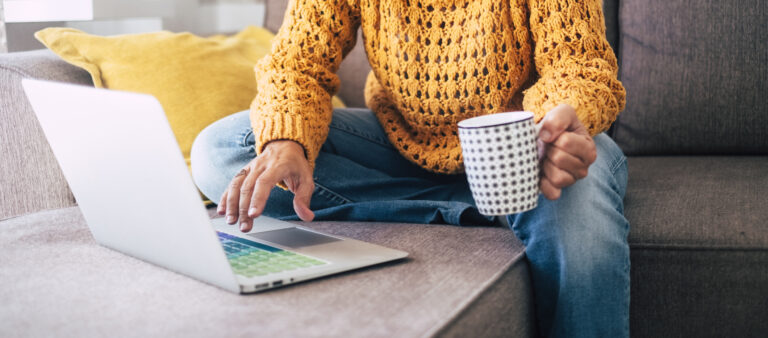 Middle aged lady with blonde curly hair and an burnt yellow sweater works at the her computer on the sofa at home