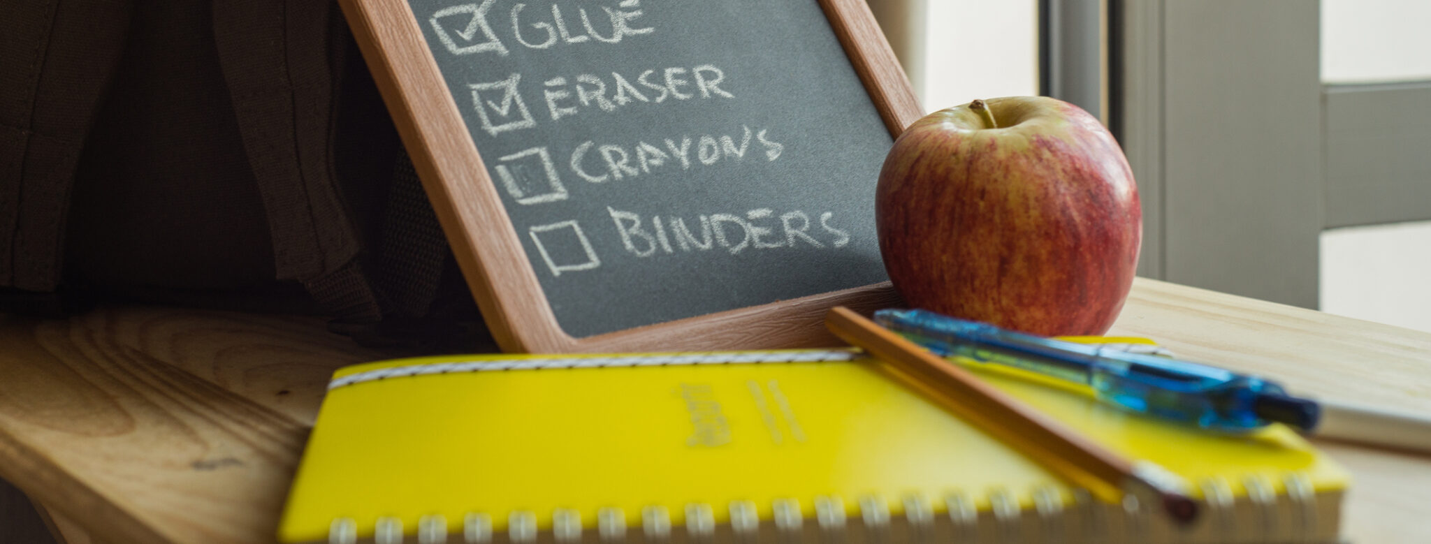 A list of school supplies on a blackboard surrounded by some school supplies. There is a backpack, notebook, pen and pencil. There is also an apple.